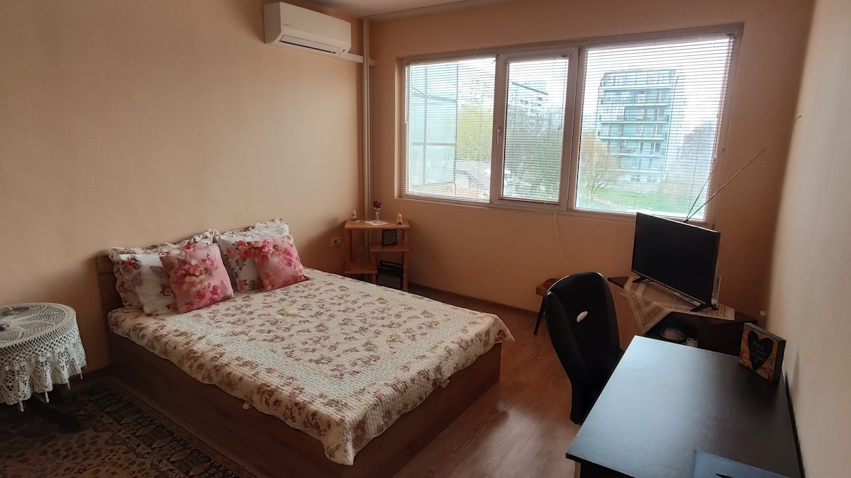 The bedroom features a spacious bed dressed in a floral quilt, complemented by decorative pillows. Large windows allow natural light to illuminate the room, and a small desk with a chair is situated alongside a television. A bedside table adds functionality to the space.