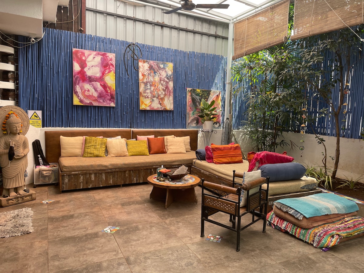 A communal lounge area features a large, cushioned sofa adorned with colorful throw pillows. The space includes decorative wall art, plants, and a low coffee table surrounded by various seating options, creating an inviting environment for relaxation and socializing.