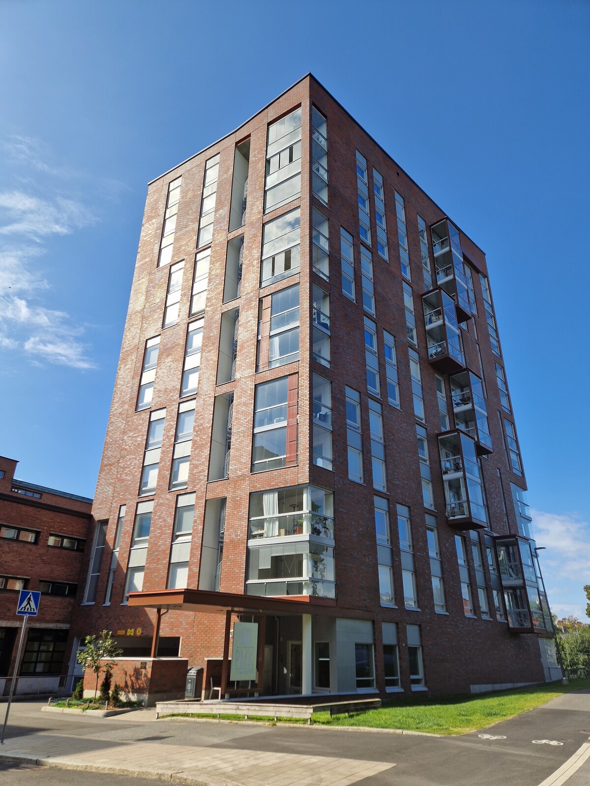 A modern brick building rises six stories, featuring a façade of large windows that reflects natural light. Balconies protrude from several floors, providing outdoor space. The surrounding area includes green grass and a paved walkway, creating an accessible entrance.