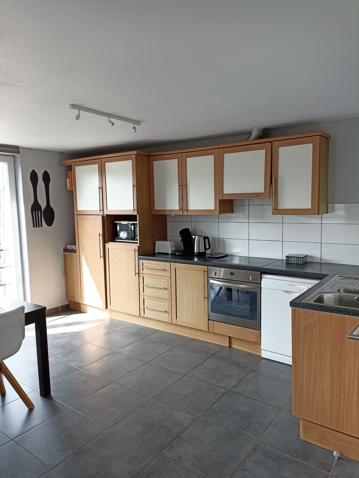 A modern kitchen features wooden cabinetry with a mix of closed and open shelving. Essential appliances, including a microwave and a dishwasher, are visible. Natural light floods the space through a sliding glass door leading to an outdoor area, with contemporary artwork displayed on the wall.