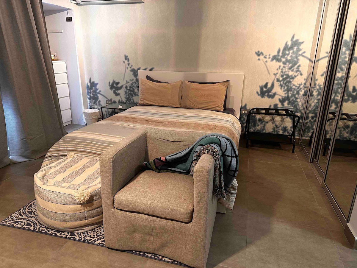 A cozy sleeping area features a queen bed with two pillows and light-colored bedding. A comfortable armchair sits nearby, adorned with a textured throw. The room displays natural shadows from nearby greenery, adding a serene ambiance to the space.