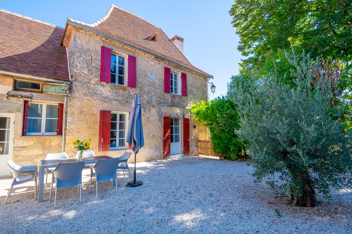 The exterior showcases a renovated stone cottage with red shutters and a gravel courtyard. A dining set with chairs and a table is arranged under the shade of a large tree, creating an inviting space for outdoor meals.