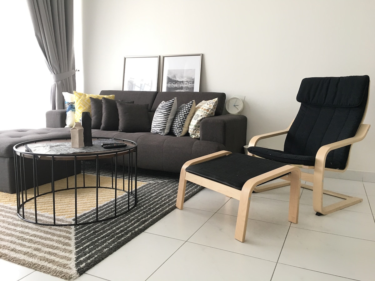 The living area features an L-shaped sofa adorned with patterned cushions and a minimalist coffee table. A stylish armchair sits adjacent, with light filtering through the sheer curtains and a neutral-toned rug anchoring the space.