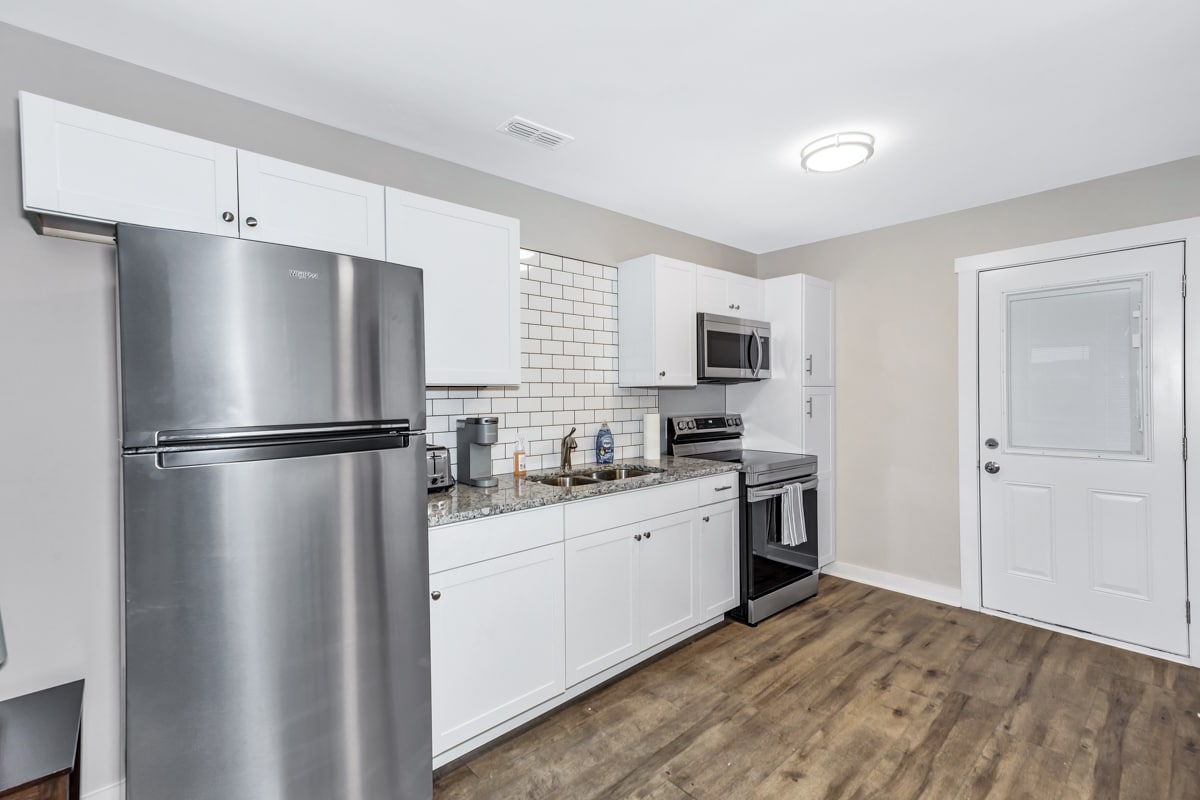 A modern kitchen is presented, featuring stainless steel appliances, including a refrigerator and oven. White cabinets complement the granite countertops. A tile backsplash adds subtle detail. Natural light is provided by a nearby door, leading outside.