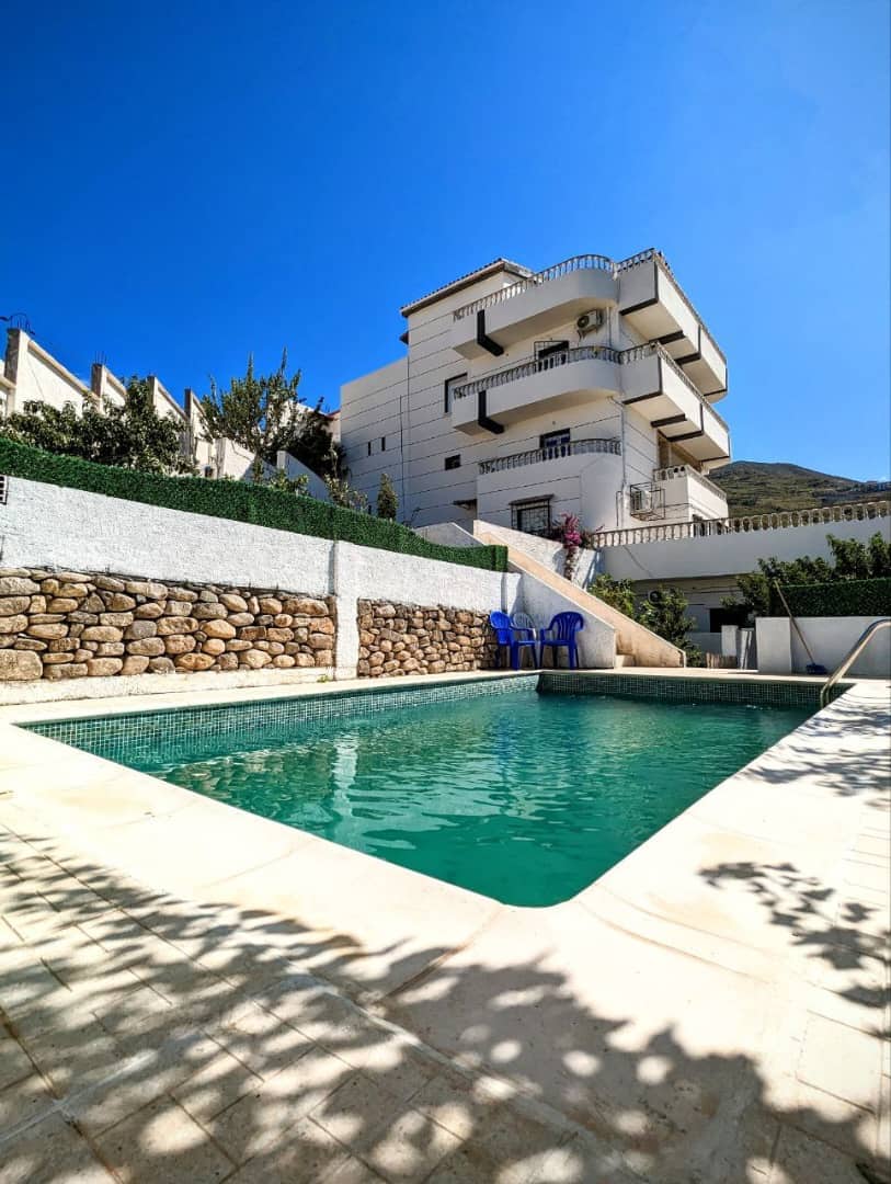 A clear swimming pool is framed by a stone wall and surrounded by greenery. Two blue chairs are positioned beside the pool. The three-story villa rises above, featuring balconies and air conditioning units, set against a bright blue sky.