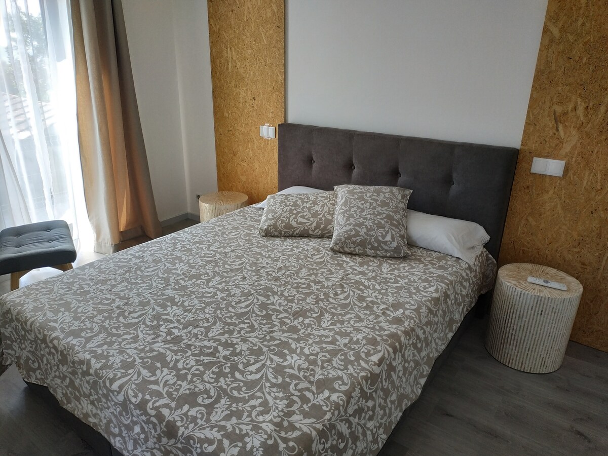 A spacious bed is outfitted with a patterned coverlet and plush pillows. Light-colored curtains frame a window, allowing natural light to filter into the room. The modern design is complemented by two round side tables and an accent chair placed nearby.