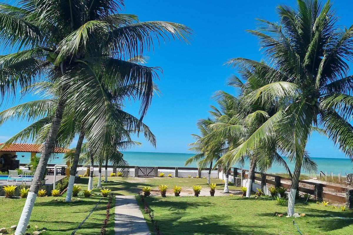 Lush green palms line a well-maintained pathway leading to the beach, with clear blue skies above. The serene ocean is visible in the background, complementing the vibrant greenery and colorful landscaping along the pathway.