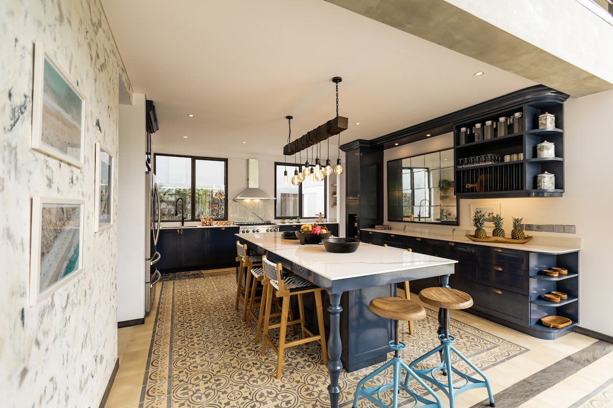 A spacious kitchen is designed with a central island and bar stools. Dark cabinetry complements the light countertops. Natural light floods through large windows, highlighting decorative shelving and modern appliances. Intricate tile flooring adds a distinctive touch to the overall aesthetic.