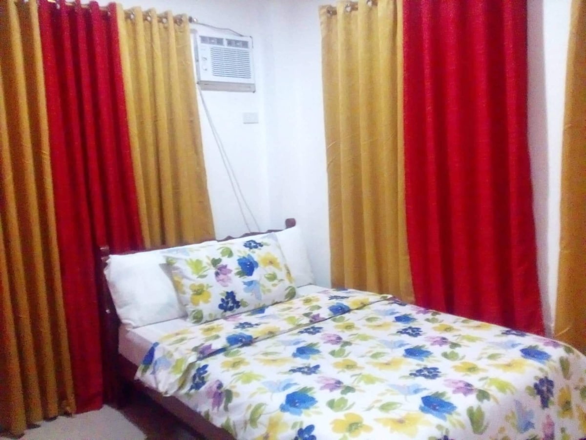 A well-decorated sleeping area features a spacious bed adorned with a floral patterned blanket and matching pillows. Bright curtains in red and gold frame the window, while an air conditioning unit is positioned above for climate control.