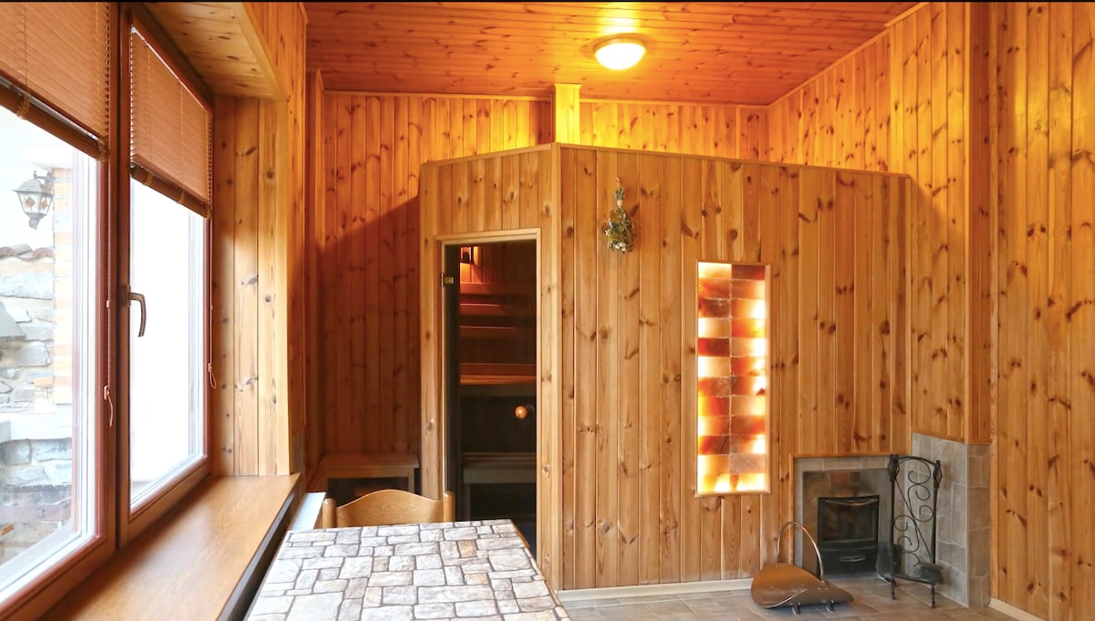 A wooden sauna is featured in a cozy room, surrounded by natural wood paneling that adds warmth to the space. Two large windows allow natural light to enter, while a stone fireplace offers an inviting focal point. A small table is visible along one wall.