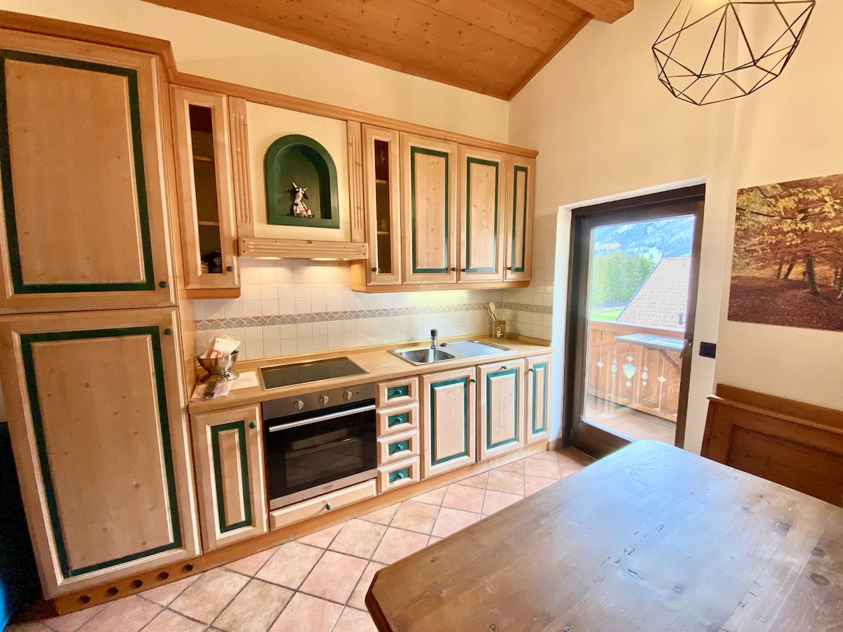 A well-equipped kitchen features wooden cabinetry with green accents, seamlessly blending rustic charm with functionality. Modern appliances are integrated, and a window offers views of the outdoors. A wooden dining table complements the space, creating an inviting area for meal preparation and enjoyment.