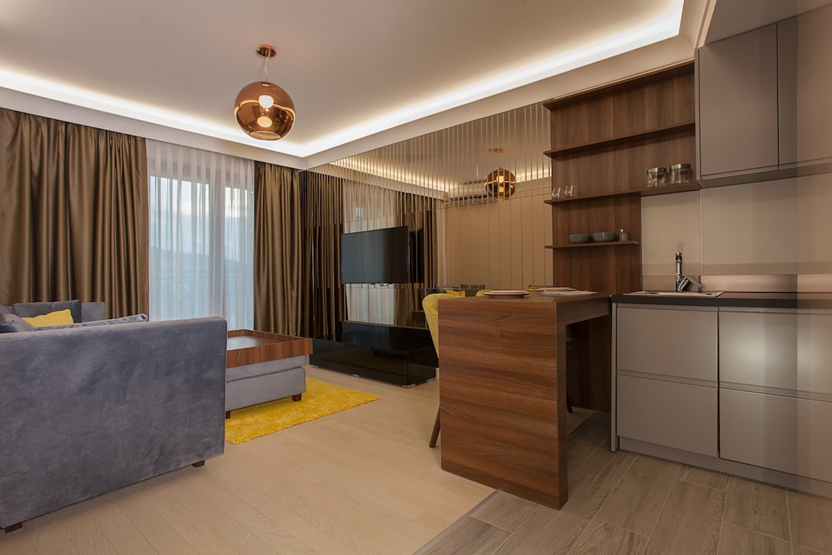 A modern living space features a comfortable blue sofa paired with a wooden coffee table. A sleek kitchen area is visible, equipped with contemporary cabinetry and a small dining arrangement. Natural light floods the room, highlighting the elegant decor and soft, neutral tones.
