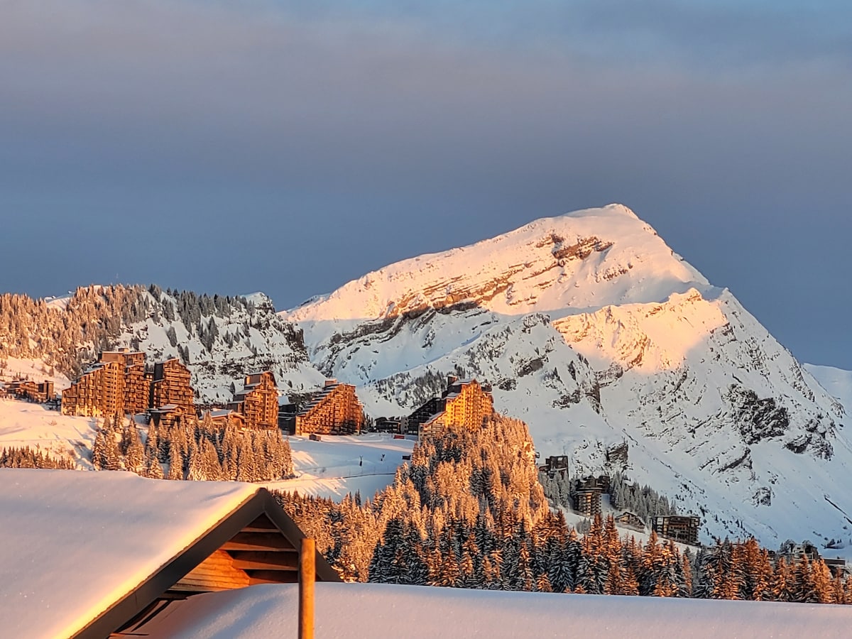 The image shows a snow-covered mountainous landscape at sunrise. Sunlight bathes the peaks and buildings in warm hues, contrasting with the cool white snow. The background features a large mountain, while the foreground includes a wooden structure, possibly a roof, highlighting the region's alpine charm.