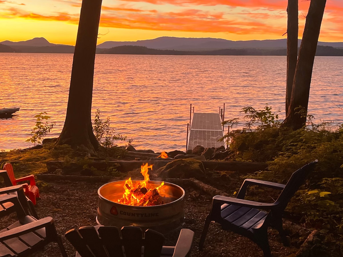 A fire pit is surrounded by several chairs, casting a cozy glow as flames dance. The backdrop features a serene lakeside view, with vibrant sunset colors reflecting on the water and mountains in the distance, creating a tranquil atmosphere.