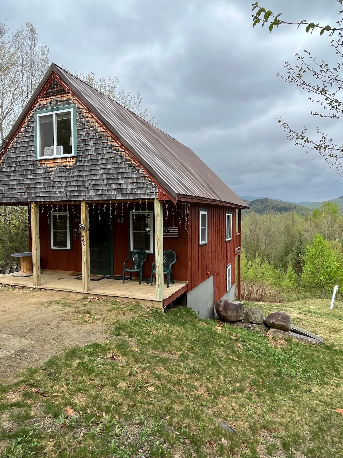 The exterior features a red cabin with a sloped roof, situated on a gentle slope. A small porch with two green chairs provides a welcoming entry. Surrounding trees and hills create a lush backdrop under a cloudy sky.
