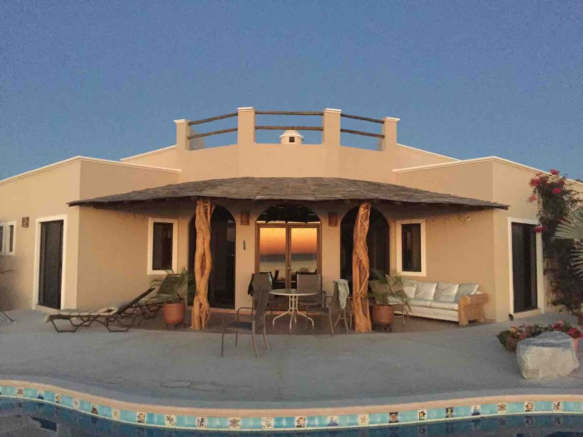 The exterior of a classic Mexican home is showcased at dusk, featuring a spacious terrace with a thatched roof. Lounge chairs and dining furniture are arranged near a sparkling pool, with panoramic views of the Sea of Cortez visible through expansive glass doors.