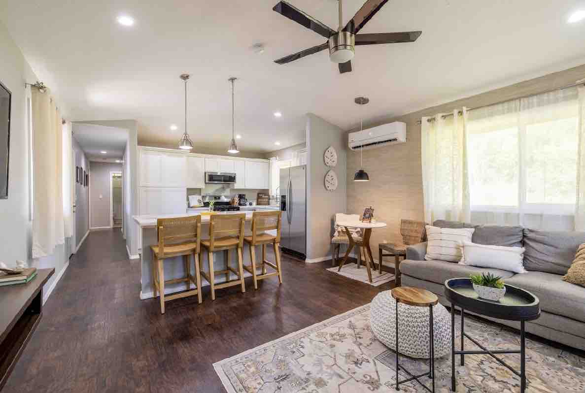 The open living area features a modern kitchen with white cabinetry and stainless steel appliances, seamlessly connecting to a dining space with bar stools. A cozy seating area is arranged with a comfortable sofa and decorative accents, illuminated by natural light from large windows.