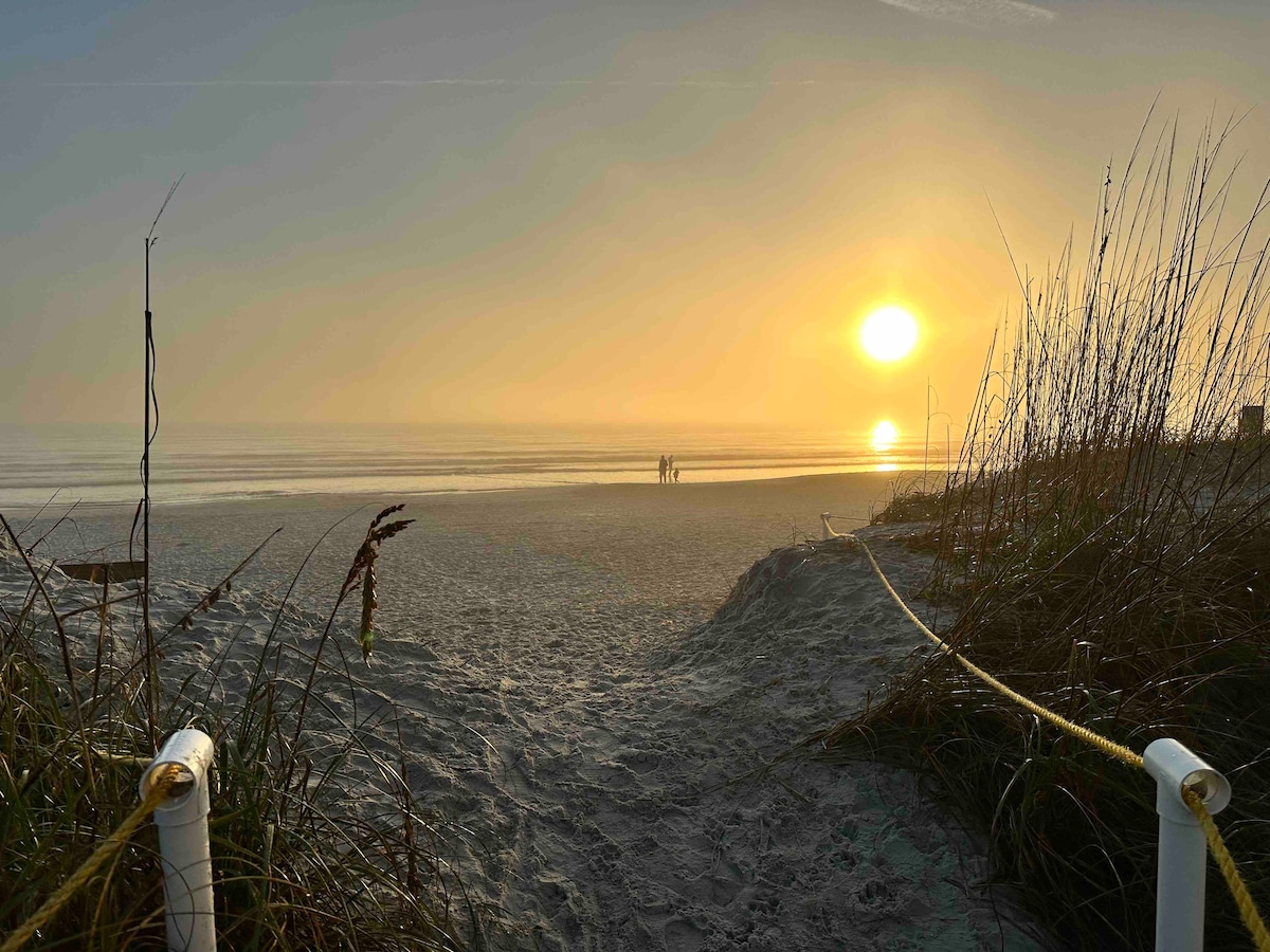 A serene beach scene captures the sun rising over the ocean, surrounded by tranquil sand and grass. Two figures are seen walking along the water's edge, with gentle waves lapping at the shore, creating a peaceful atmosphere.
