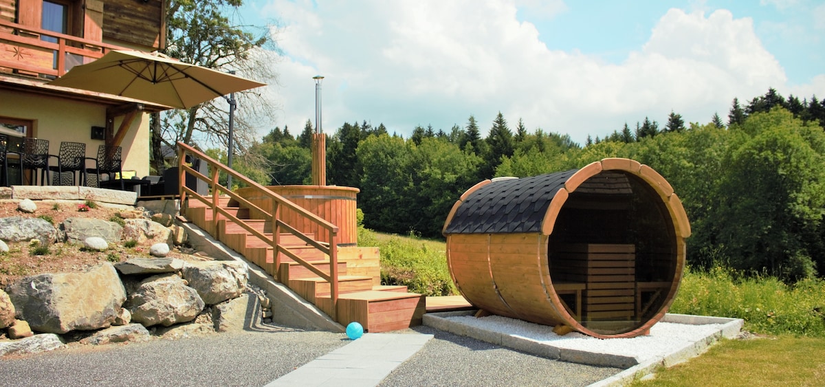 An outdoor area features a wooden sauna designed in a barrel shape, surrounded by greenery. Steps lead from the building to the sauna, ensuring accessibility. A large umbrella provides shade over a wooden seating area with several chairs visible. A pebble path runs along the side.