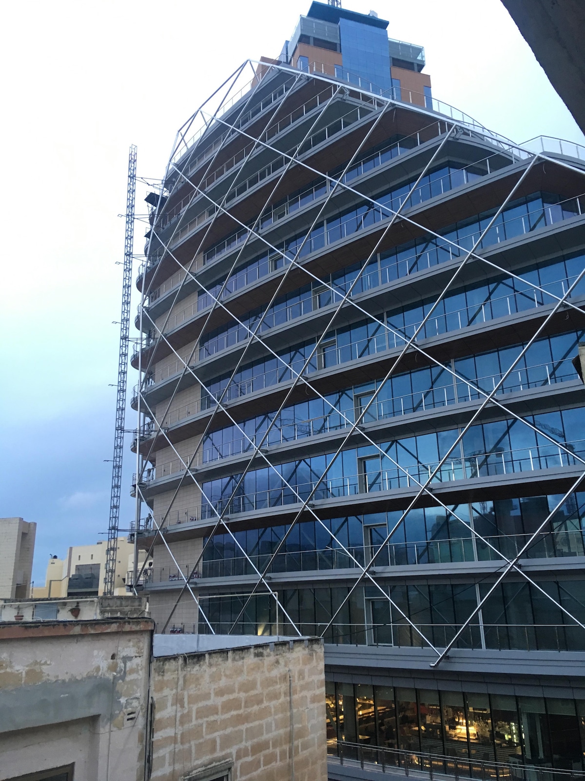A modern building features a unique architectural design with an angled facade and large glass windows. The structure's exterior is enhanced by diagonal metal supports, reflecting contemporary style. The surrounding area includes lower buildings, creating a contrast in height and design.