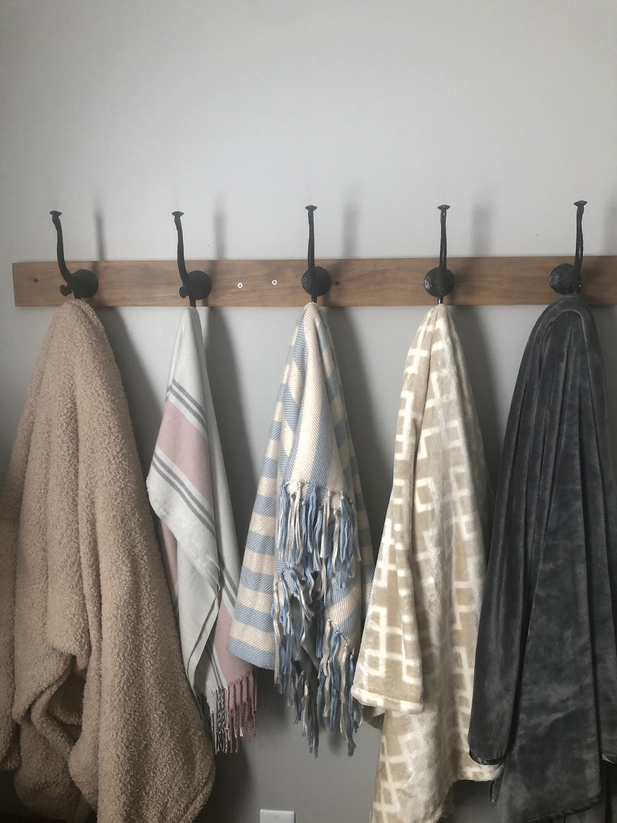 A wooden wall-mounted rack holds five different blankets. Varieties include a soft beige throw, a light grey with a geometric pattern, a pastel striped blanket, and a checkered blue and white blanket. The arrangement provides easy access to cozy coverings.
