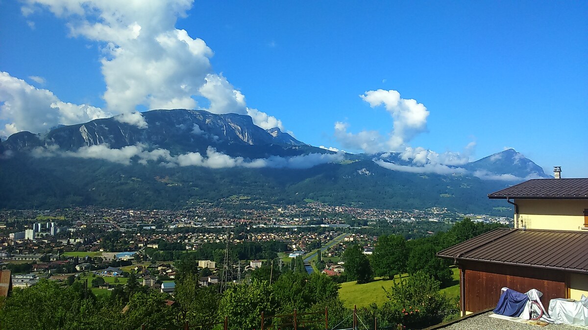 A scenic view of mountains and valleys is captured, with rolling hills leading down to a town. Partly cloudy skies are visible above, illuminating the landscape below. The lush greenery and distant structures hint at a peaceful rural environment.