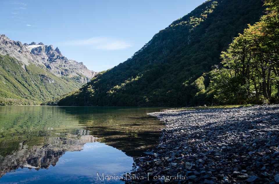 The serene landscape features a calm lake reflecting the surrounding mountains and forest. Pebbles line the shore, with lush greenery rising from the water's edge. The clear sky creates a tranquil atmosphere, highlighting the natural beauty of the area.