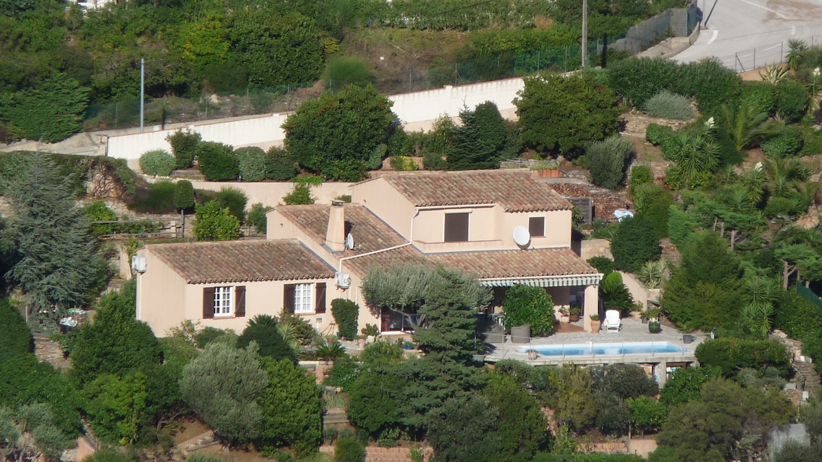 An aerial view of a single-family villa surrounded by lush greenery. The property features a tiled roof, multiple windows, and an outdoor pool. The landscaping includes various trees and shrubs, providing a serene atmosphere. A terrace extends along the villa, enhancing outdoor living space.