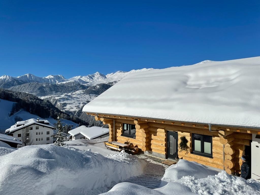 A newly built log cabin is nestled among snow-covered terrain, featuring a sloped roof and large windows. The surrounding mountains create a serene backdrop, while the wooden exterior adds rustic charm. Snow blankets the ground, enhancing the cabin's winter setting.