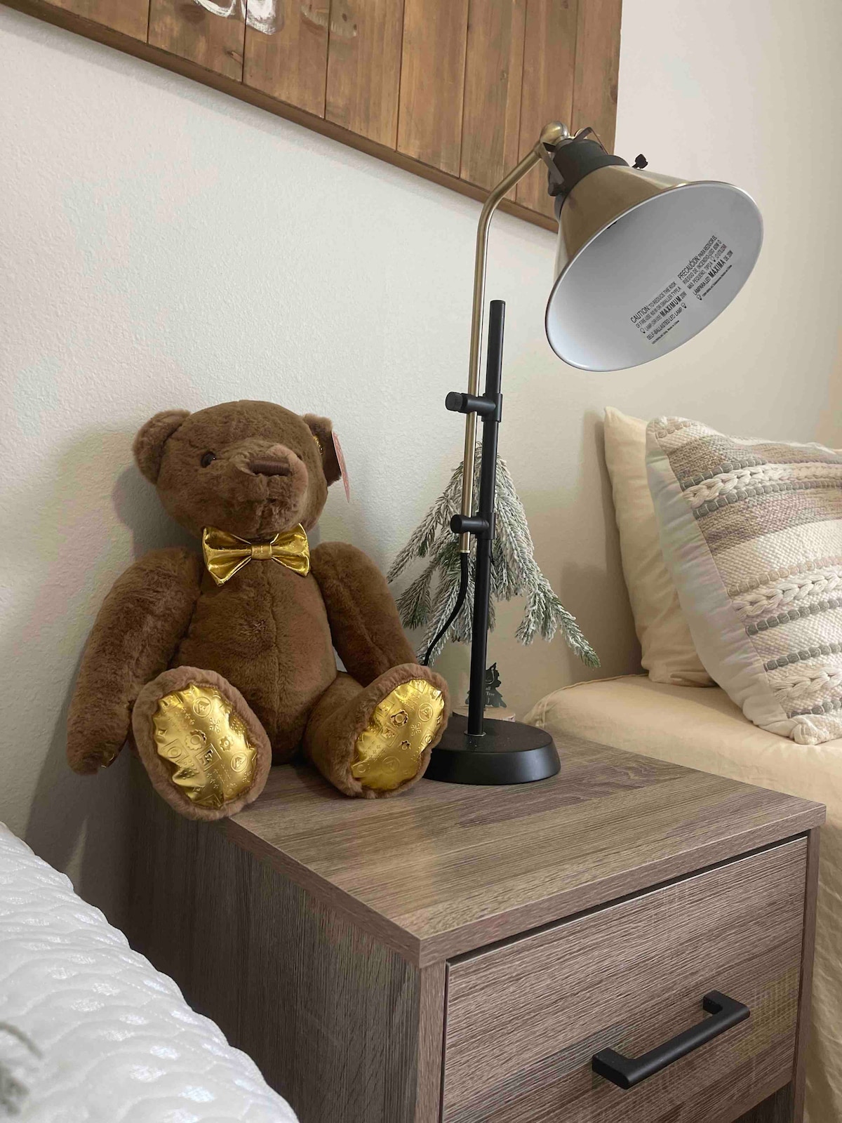 A bedside table is featured, supporting a stylish lamp and a plush teddy bear with a golden bow. Soft, textured pillows are arranged on the bed nearby, complementing the warm tones of the room's decor.