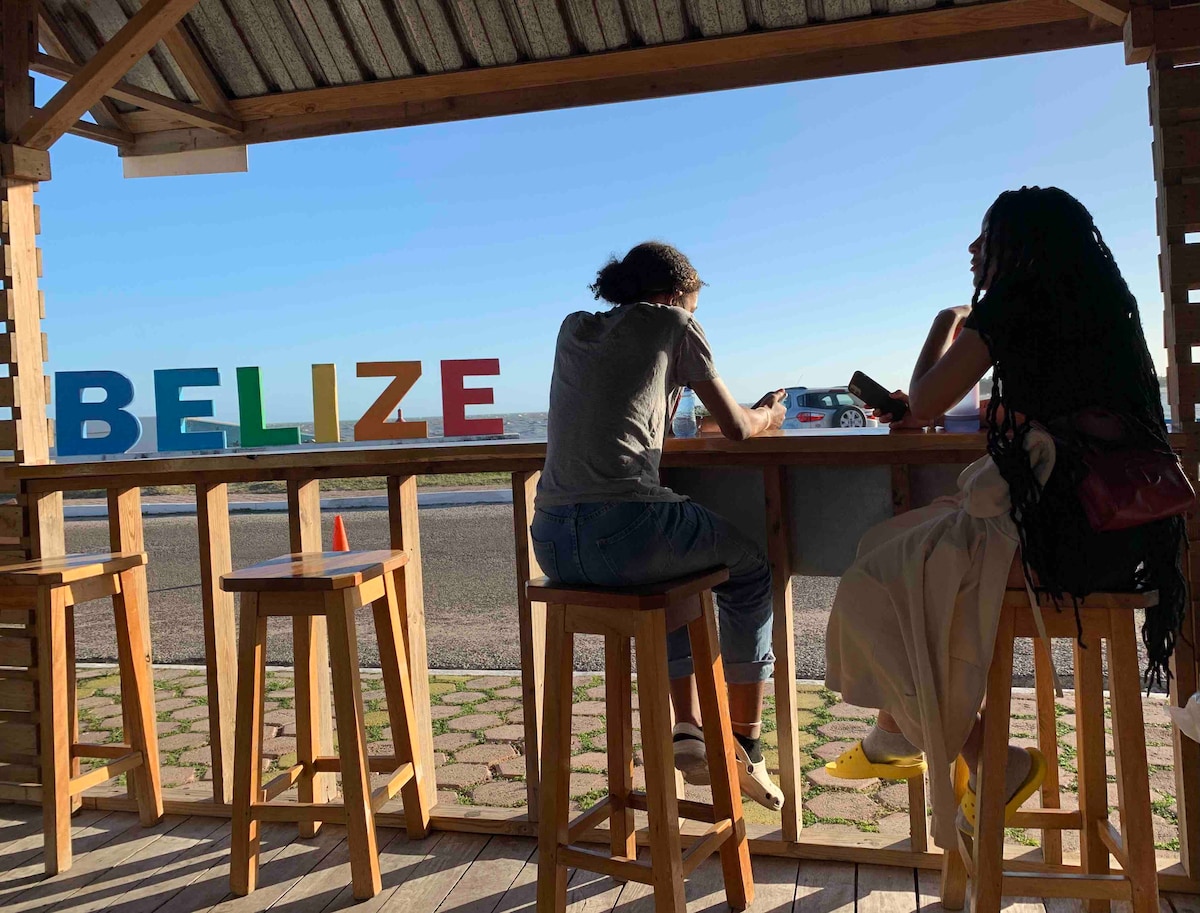 A wooden bar area is featured with high stools, providing views of the horizon. Colorful letters spelling 'BELIZE' are displayed prominently, adding a vibrant touch to the serene setting. The open space allows for natural light to illuminate the space.