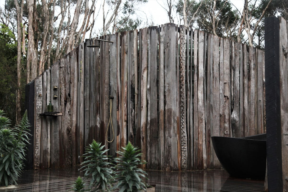 An outdoor bathing area is enclosed by a rustic timber privacy screen, providing seclusion. A sleek black stone bath is positioned beside the decking, complemented by tall, lush plants that enhance the tranquil environment.