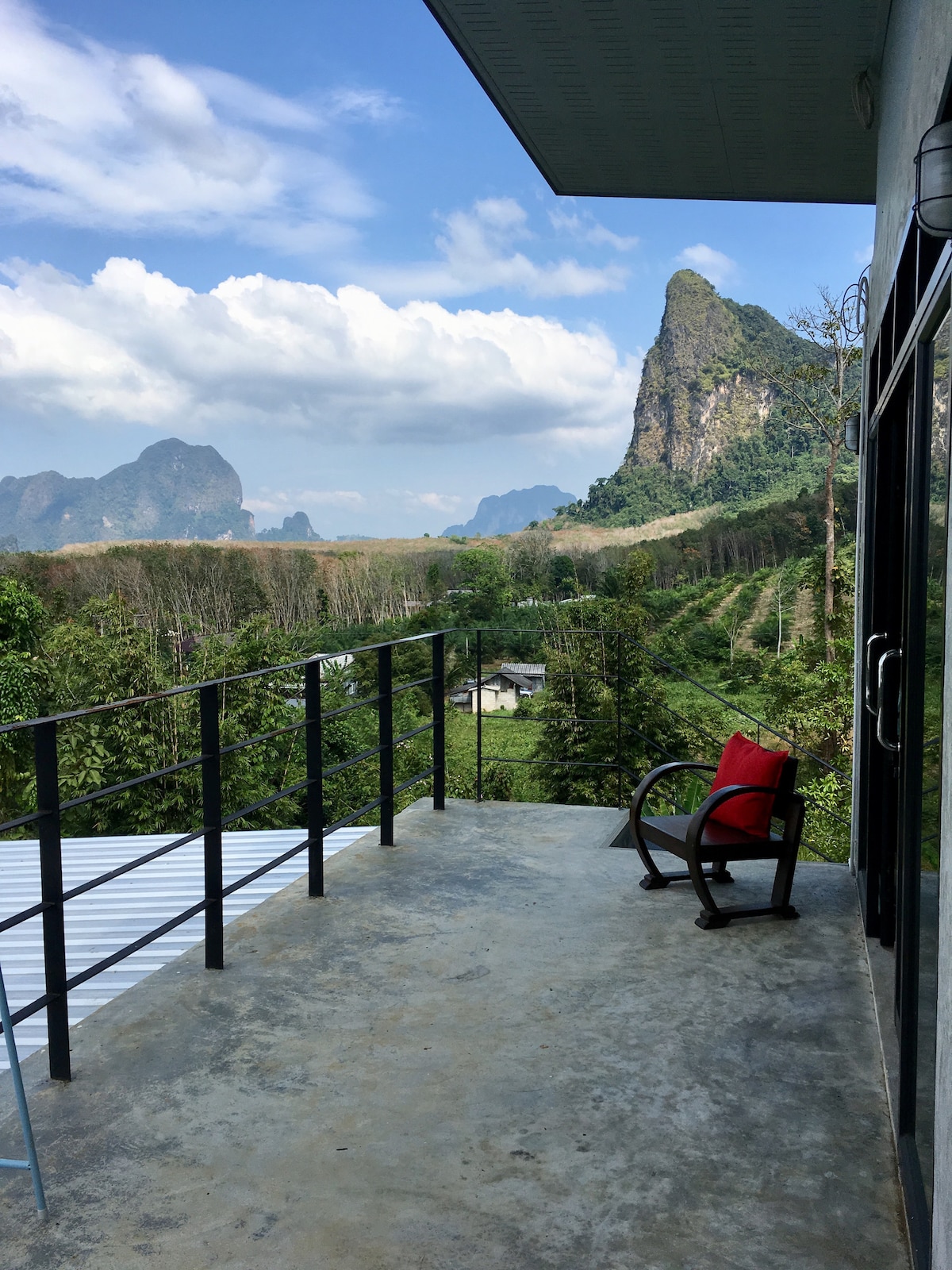 A spacious terrace features a wooden rocking chair and offers unobstructed views of the surrounding mountains and lush greenery. The sky is partly cloudy, and the landscape is layered with hills, creating a serene atmosphere ideal for relaxation.