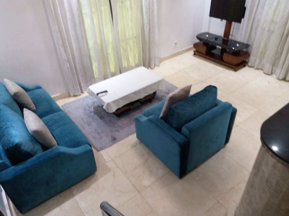 A bright living area is furnished with two teal sofas, accompanied by decorative pillows. A white coffee table rests on a soft gray rug, while a television is mounted on a dark wooden stand nearby. Sheer curtains allow natural light to filter in.