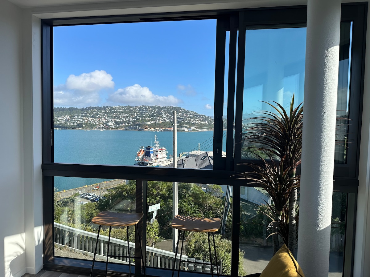 A large glass window showcases scenic views of Wellington Harbour, with a ship visible in the foreground. Two wooden bar stools are positioned by the window, allowing for an inviting space to enjoy the natural light and the picturesque surroundings.