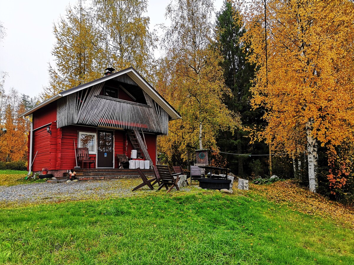 A two-story red cottage is surrounded by autumn foliage, displaying vibrant yellow and orange leaves. A wooden staircase leads to the entrance, and patio chairs are positioned on the front terrace. A fire pit is visible in the garden, inviting moments of relaxation outdoors.