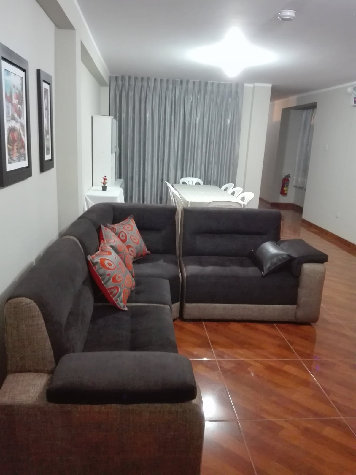 A spacious living area is furnished with a dark sectional sofa, featuring decorative pillows for comfort. A dining table with white chairs is visible in the background, complemented by large windows covered with light gray curtains. The polished floor reflects the room's lighting.