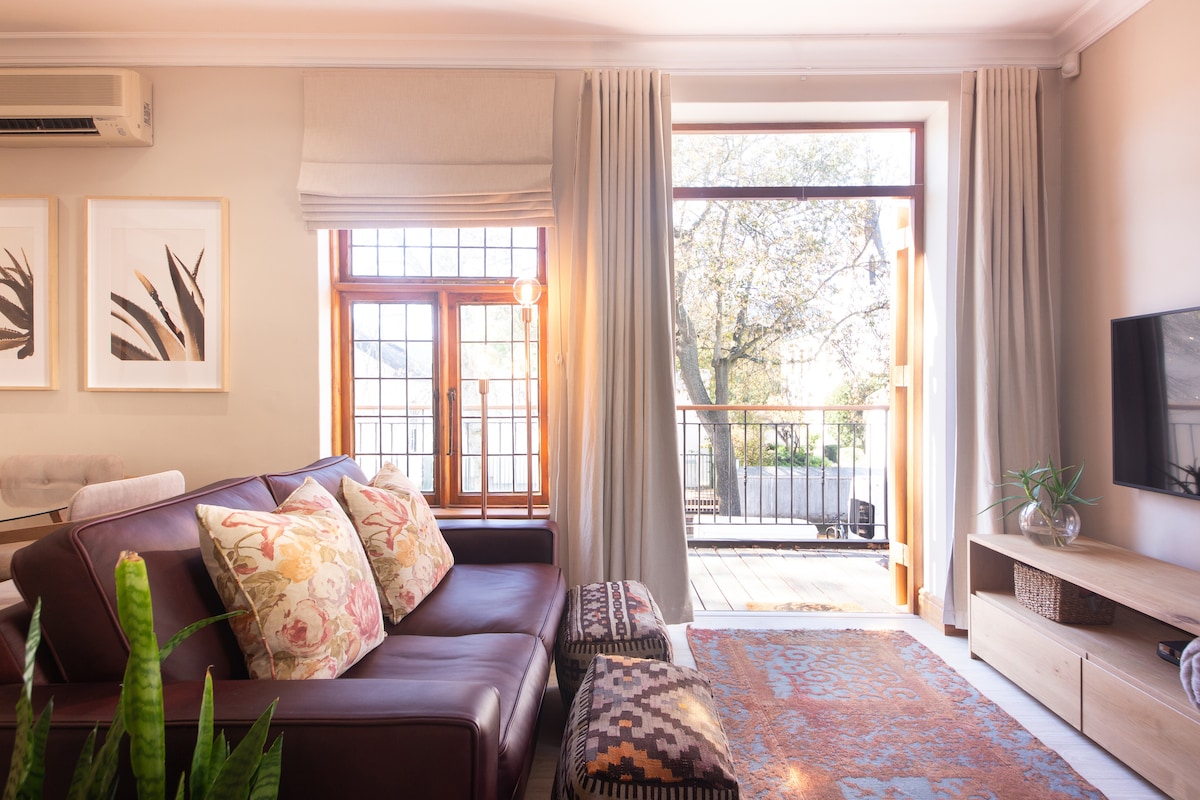 A sunlit living area features a comfortable leather sofa adorned with floral cushions. Large windows frame views of the balcony and greenery outside, with sheer curtains allowing soft light to filter in. A wooden media console sits beside a flat-screen TV, enhancing the space's function.