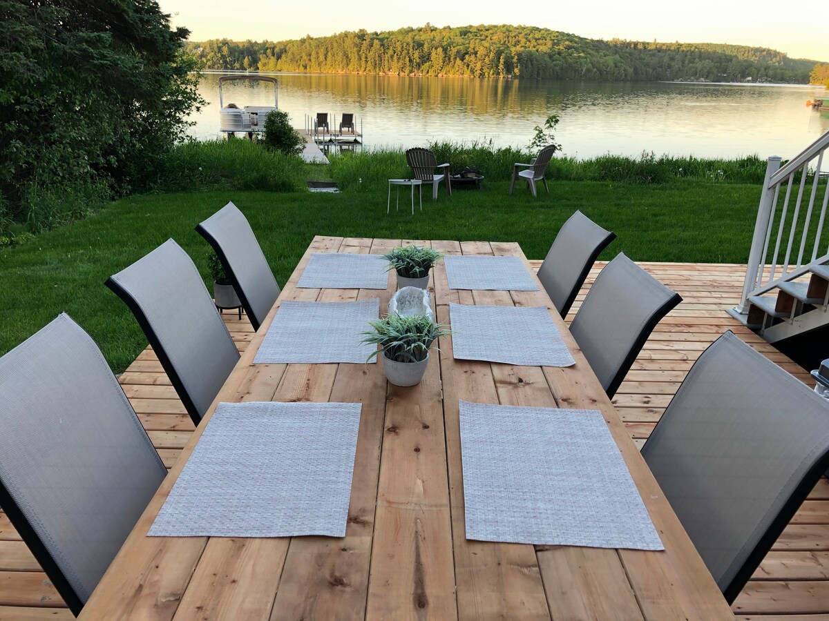 A large wooden dining table is set outdoors with gray placemats and surrounding chairs. Lush greenery and a scenic view of the lake are visible in the background, featuring calm waters and distant trees. A small dock can be seen along the shoreline.