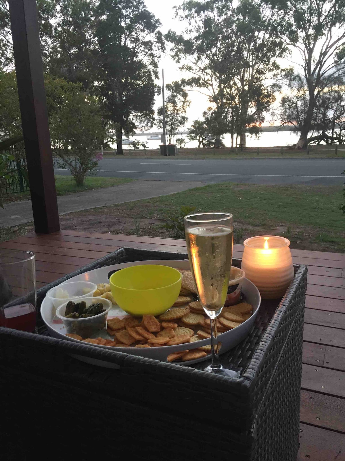 A tray of snacks, including crackers, olives, and sliced lemons, is set on a wicker table. A glass of sparkling drink rests prominently beside a lit candle, with tranquil water views and trees in the background, highlighting a relaxed outdoor setting.