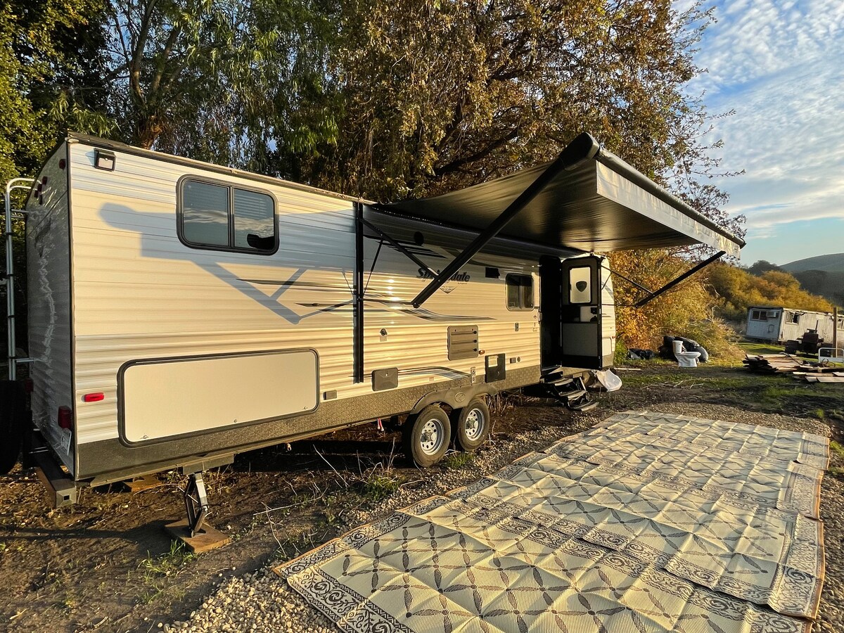 A well-maintained camper is positioned under a blue sky, featuring an extended awning for shade. A patterned outdoor rug is laid out outside the camper, creating an inviting space for relaxation. Nearby, the natural landscape of the Milpitas Mountains is visible.