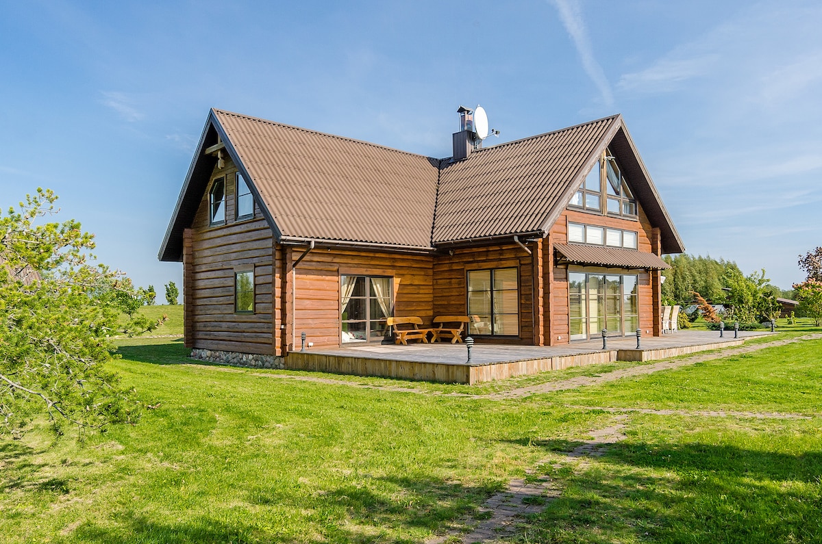 The wooden country house features a spacious porch with outdoor seating. Large glass windows allow natural light to fill the interior. Surrounding greenery enhances the scenic view. The structure is framed by a well-maintained lawn and is set against a clear blue sky.