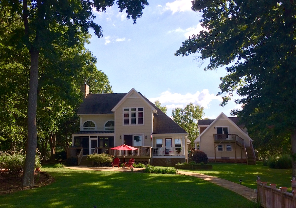 A charming waterfront property is showcased, featuring a main house with large windows and a welcoming porch. A smaller structure stands nearby, and a circular pathway is seen in the foreground, complemented by a red patio umbrella and seating area surrounded by lush greenery.