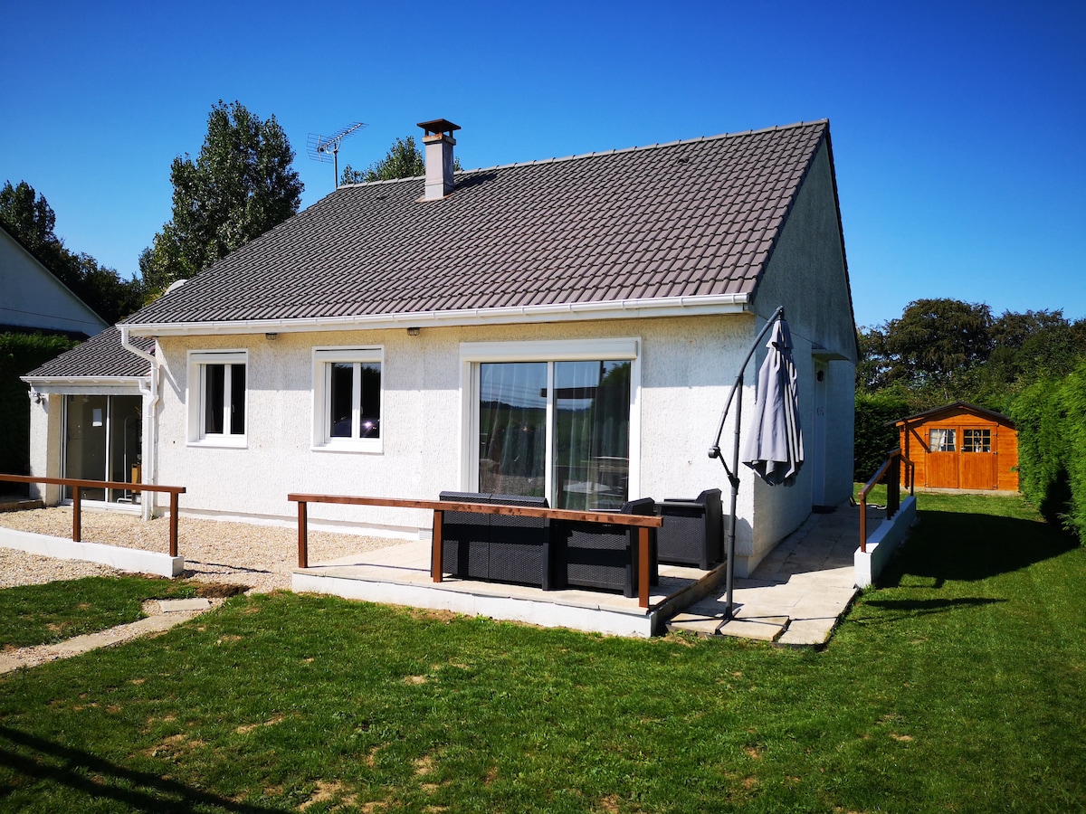 A single-story house is set against a clear blue sky, featuring a gray tiled roof and light exterior walls. A spacious patio with a seating area overlooks the green lawn. A wooden garden shed is visible in the background, surrounded by trees.