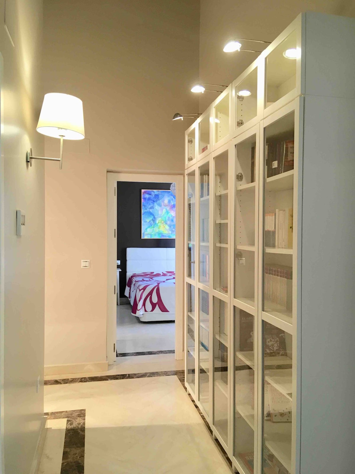 A well-lit hallway is lined with a tall bookshelf featuring glass-fronted compartments, displaying various books and decorative items. A soft lamp on the wall provides additional lighting. At the end of the hallway, a doorway leads to a bedroom with vibrant bedding and colorful artwork.