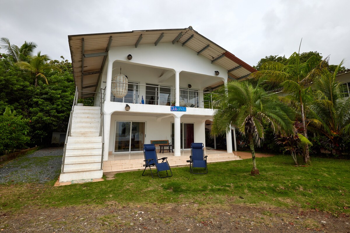 A two-story house is positioned against a lush green backdrop. A spacious porch features garden chairs overlooking a grassy area. The exterior highlights a mix of modern design and natural surroundings, with large windows allowing for ample natural light.