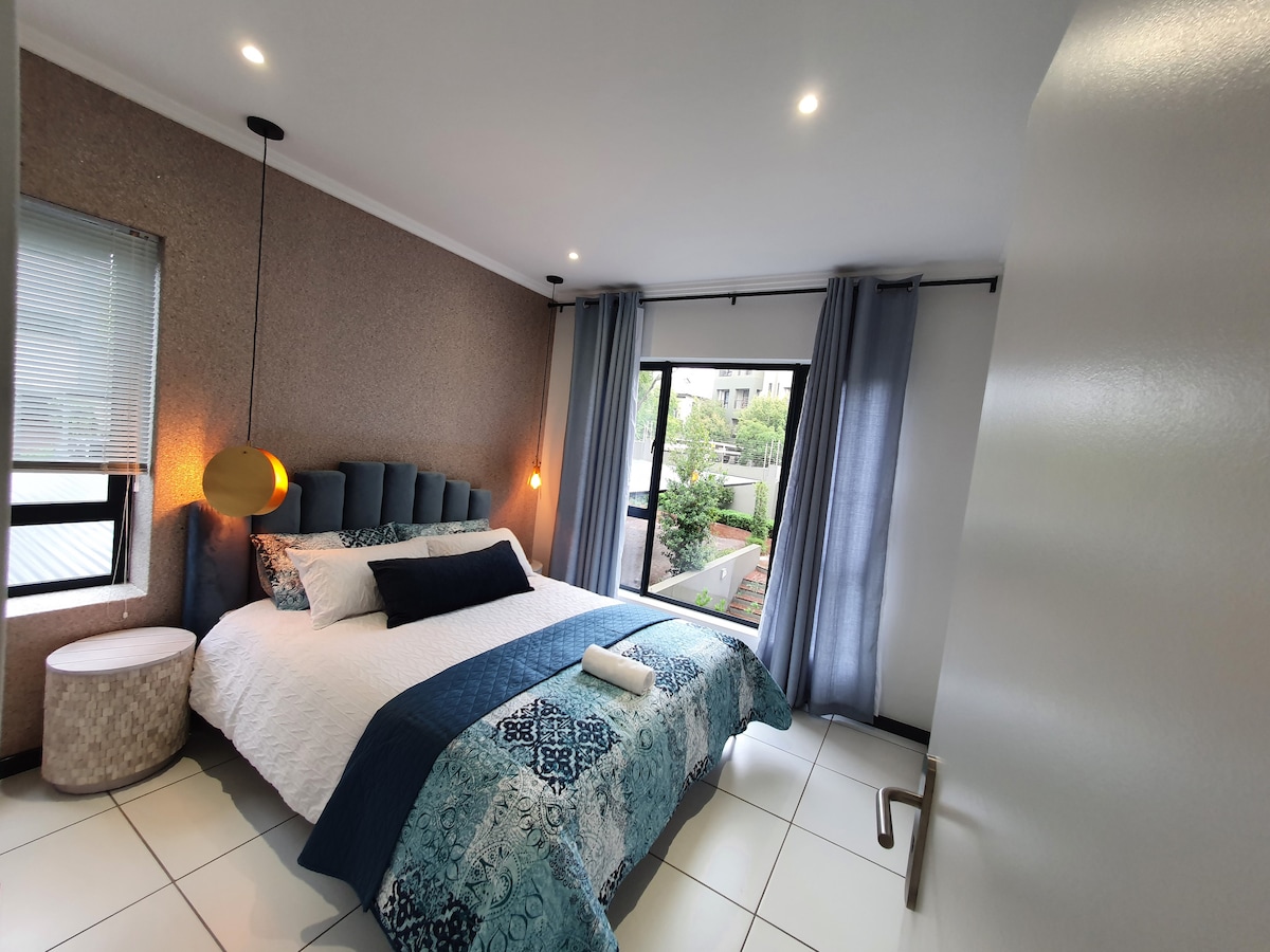 A spacious bedroom features a large bed with a patterned quilt and soft pillows. Natural light enters through windows, framed by sheer curtains, with a cozy side table beside the bed. Modern decor is complemented by a subtle color palette.