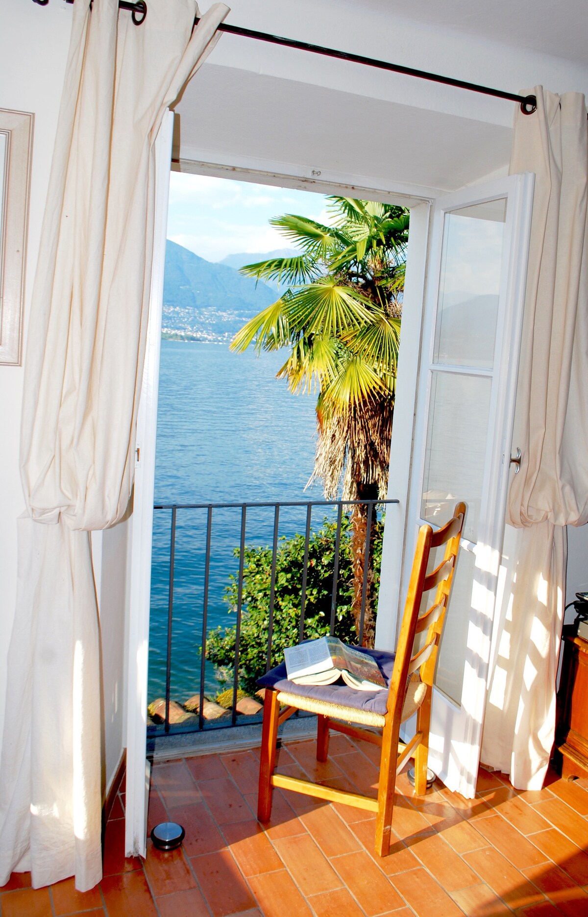A wooden chair is positioned next to an open window, providing a view of palm trees and Lago Maggiore. Natural light streams in through the white curtains, highlighting the warm tones of the terracotta flooring.