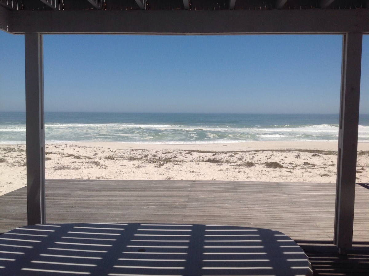 A view of the Atlantic Ocean is framed by a covered outdoor area, highlighting a sandy beach that stretches along the shoreline. Soft waves gently lap at the shore under a clear blue sky, creating a serene coastal atmosphere.