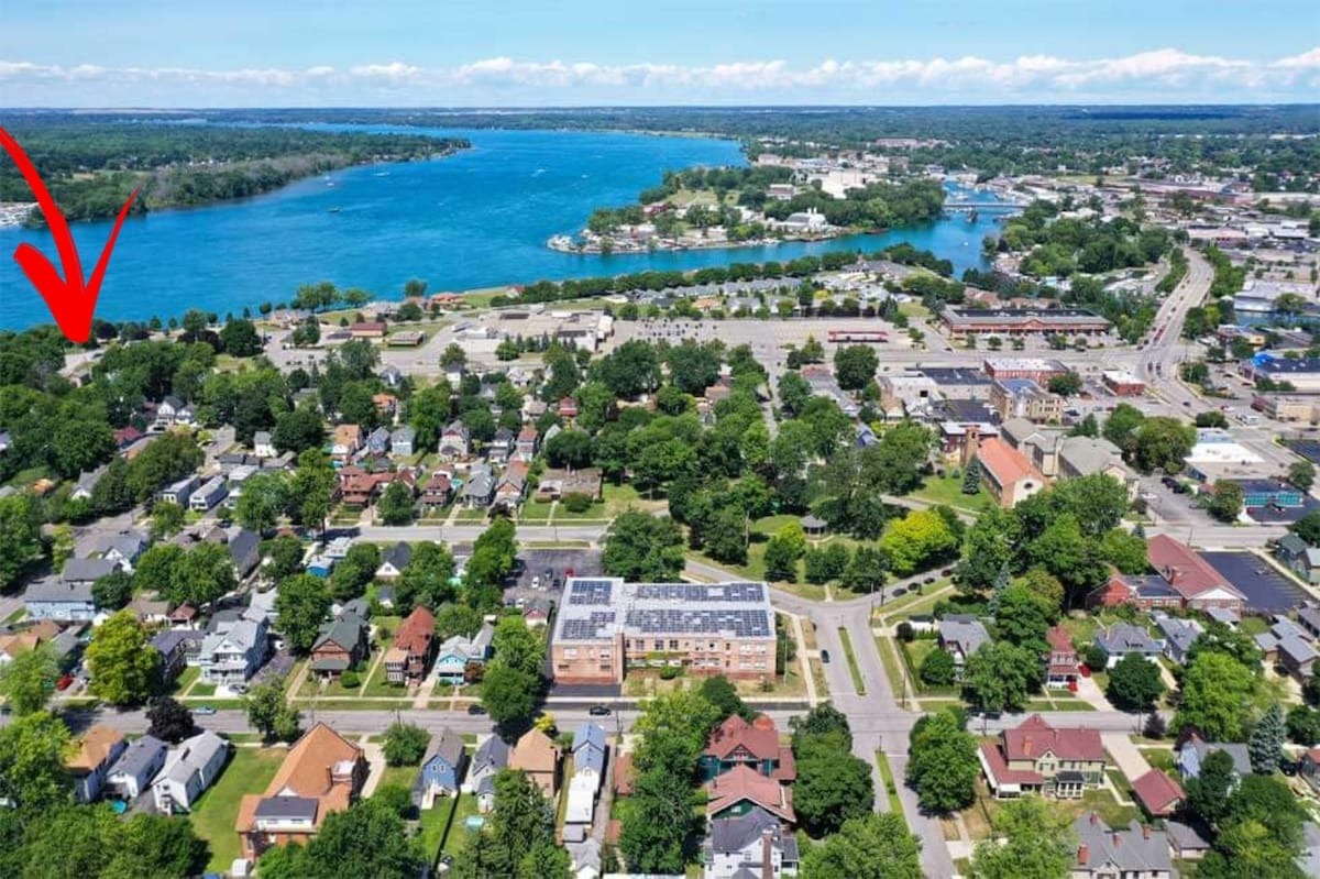 An aerial view captures a charming neighborhood bordered by the Niagara River, showcasing lush greenery and residential homes. The landscape features winding waterways, with the yurt location indicated near the vibrant, blue waters. Scenic parks and local amenities are visible in the surrounding area.